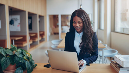 Young African American businesswoman smiling while standing at a table in the lounge of a modern office working on her laptop